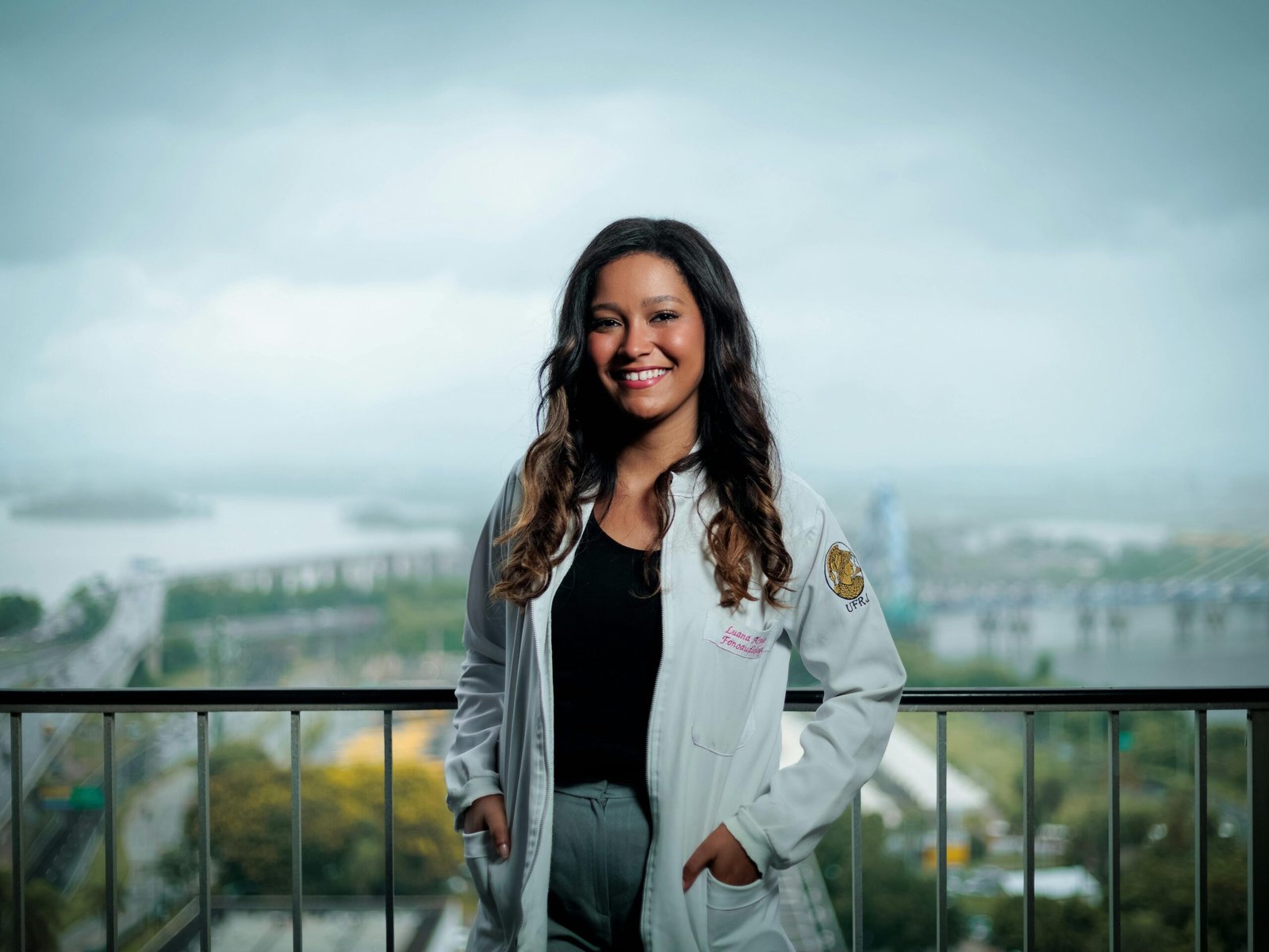 Smiling female doctor in white coat posing on a balcony with a cityscape background.